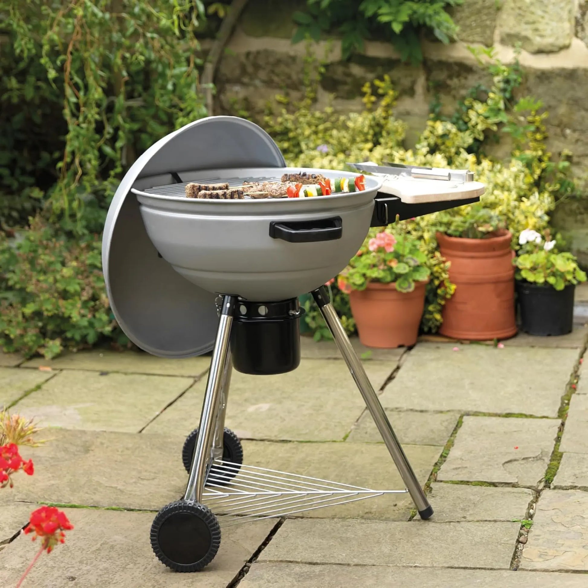 Barbecue grill on wheels with food cooking outdoors, surrounded by potted plants.