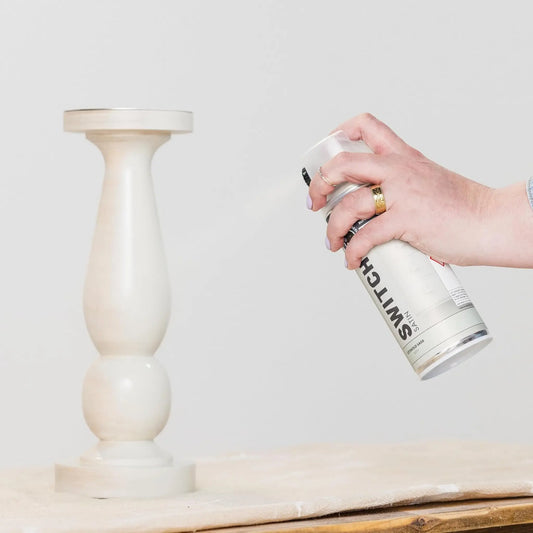 Person spray painting a wooden candle holder with a Switche brand can on a light background